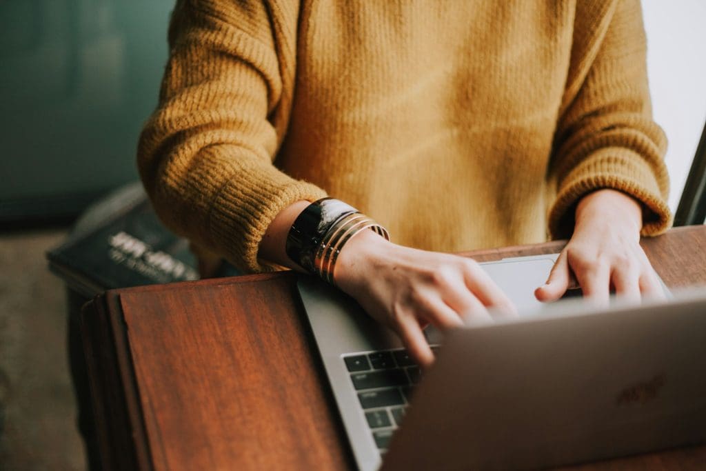 hands and torso of person in mustard colour sweater on laptop resting on a wooden desk in daytime