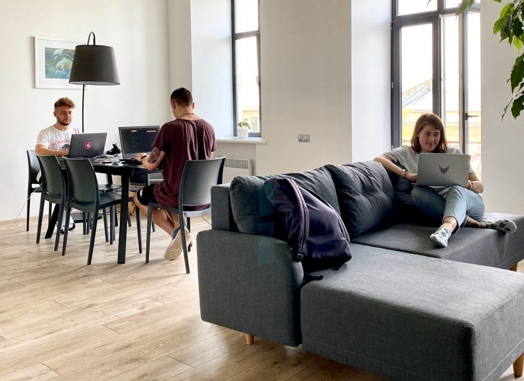 A woman sat on a sofa with a laptop and two men sat at a dining table with two laptops